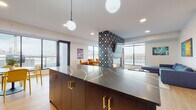 This single-floor space showcases a thoughtfully designed open-plan layout that seamlessly connects multiple functional zones, including a kitchen, lounge, and dining areas. The kitchen is anchored by a striking dark-toned island with bold veining detail and brushed gold hardware, complemented by dark subway tile backsplash, matte cabinetry, and globe pendant lighting overhead. Blue velvet barstools line the island, adding a pop of color against the moody palette.

A dramatic floor-to-ceiling tiled column serves as the space's visual centerpiece, clad in a bold geometric pattern in deep navy, teal, and white, and housing a linear fireplace. Adjacent to the fireplace, the lounge area features a generous sectional sofa in steel blue, a circular coffee table, and vibrant large-format artwork adorning the walls. Warm light pours in through expansive floor-to-ceiling windows on multiple sides, offering sweeping urban views and filling the space with natural light.

A separate seating nook furnished with rust and amber velvet chairs provides an intimate gathering spot, while a banquette dining area with vivid yellow seating adds a playful contrast. Light wood flooring runs throughout, tying the varied zones together into a cohesive, contemporary aesthetic.