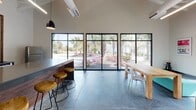 This contemporary single-story space showcases an open, industrial-inspired design with soaring exposed roof trusses and polished concrete floors throughout. The main lounge area centers around a striking double-sided fireplace clad in warm wood paneling with a concrete hearth, topped by a wall-mounted TV — a focal point that anchors the entire room. Modular gray sectional seating and patterned area rugs add color and comfort, while floor-to-ceiling black-framed windows flood the space with natural light and frame views of mature palm trees and lush landscaping outside.

A dedicated dining and bar area features a solid wood communal table, counter-height bar seating with wood stools, and expansive grid windows that open the room to the surrounding greenery. The kitchenette is outfitted with white cabinetry, dark countertops, a geometric tile backsplash, a sink, and a compact refrigerator.

A separate recreation area provides additional open space, highlighted by a pool table, exposed ductwork, and access to a glass-walled private room. Linear LED lighting runs throughout, reinforcing the clean, modern aesthetic. The overall ambiance is inviting yet polished, ideal for collaborative or social use.