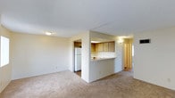 Located at 1051-1057 Broadway in Chula Vista, this Plan B unit offers a practical and well-organized single-floor layout designed for comfortable everyday living. The open-concept living area is finished with wall-to-wall carpeting, creating a warm and inviting atmosphere throughout the main space. A defining feature of this unit is the kitchen pass-through, which connects the kitchen to the living area, enhancing the sense of openness and allowing for easy interaction between spaces. The kitchen is equipped with warm-toned wood cabinetry providing ample storage, a white refrigerator, and a countertop that doubles as a casual dining or breakfast bar surface. The kitchen flooring transitions to a hard surface, offering a practical distinction from the carpeted living areas. The neutral color palette of cream and beige tones throughout the walls and finishes contributes to a bright, clean aesthetic. A hallway visible from the main living area leads to additional rooms, suggesting a thoughtful and functional floor plan. The textured ceiling and flush-mount lighting provide consistent illumination across the space. Overall, this unit presents a straightforward, move-in-ready layout well-suited for those seeking efficient and comfortable living in Chula Vista.