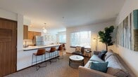 This single-floor residence offers a thoughtfully designed open-concept layout that seamlessly connects the kitchen, living, and dining areas. The kitchen is outfitted with warm wood cabinetry, white quartz countertops, stainless steel appliances, and a breakfast bar with leather-toned stools — ideal for casual dining or entertaining. Pendant lighting adds a contemporary touch overhead.

The living area is a comfortable, well-lit space featuring plush carpeting, a neutral-toned sofa accented with teal throw pillows, and a round marble-top coffee table. An arched mirror accent near the entryway and abstract wall art add character throughout. Natural light filters in through blinds on multiple windows, enhancing the bright, welcoming atmosphere.

A separate dining nook features hardwood-style flooring, a natural wood dining table with modern chairs, and botanical-framed artwork — creating an intimate setting for meals.

The bedrooms are carpeted and include ceiling fans, ample closet space with sliding doors, and windows that bring in natural light. The primary bedroom accommodates a full-size bed with warm, layered bedding and tasteful décor.

The bathroom features a wood-toned vanity with a white countertop, a large mirror, and hardwood-style flooring, offering a clean and functional design. Overall, the space presents a cohesive, modern aesthetic with warm natural wood tones balanced by crisp white walls.