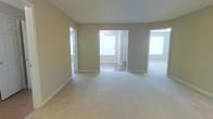 This single-story space offers a clean, neutral aesthetic defined by soft beige walls and light-colored carpeting that extends through the main living area. The open layout connects seamlessly to an adjoining room visible through a wide doorway, where hardwood or laminate flooring contrasts with the carpeted main space. That secondary room benefits from a ceiling fan and large windows that invite generous amounts of natural light, creating a bright and airy atmosphere. An additional opening to the right suggests further functional space, also well-lit by natural light. A door to the left appears to lead to an exterior area, potentially a patio or balcony, adding an outdoor connection. White baseboards and trim provide clean visual contrast against the warm neutral walls throughout. The overall ambiance is understated and versatile, with a move-in-ready feel suited to a variety of furnishing styles. The interconnected flow between rooms enhances the sense of openness, making the space feel larger than its individual components might suggest.