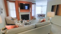 Located at 2400 Garden Hill Dr in Raleigh, North Carolina, this single-floor residence offers a thoughtfully designed layout that balances style and comfort. The living area serves as the heart of the home, anchored by a classic white-mantled fireplace set against a striking burnt-orange accent wall that adds warmth and visual depth to the space. A wall-mounted flat-screen television is positioned above the fireplace, making it a natural focal point. Plush neutral-toned seating, accented with patterned and orange throw pillows, creates an inviting and cohesive atmosphere. A set of nesting coffee tables sits at the center of the seating arrangement, adding both function and style. The living area flows seamlessly into a dedicated dining space, which features a dark wood dining table and chairs beneath a decorative pendant light fixture. Multiple windows throughout both areas are dressed with white plantation shutters and sheer patterned curtains, allowing generous natural light to fill the rooms while maintaining privacy. Neutral carpet flooring ties the spaces together, contributing to the home's warm and welcoming ambiance. The overall design combines bold color choices with classic architectural elements for a polished, livable interior.