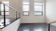 This contemporary multi-level residence spans three floors, offering a well-organized layout that balances open living areas with private retreats. The main level, the largest of the three, features a striking modern kitchen equipped with stainless steel appliances, dark cabinetry with brushed hardware, a tiled backsplash, and a quartz waterfall island with an undermount sink — all illuminated by track lighting and pendant fixtures beneath soaring ceilings. Exposed ductwork lends an industrial character to the space.

The staircase area is a design focal point, featuring bold dark-toned stairs with contrasting light treads, gold-accented handrails, cable railings, and pendant lighting that highlight the vertical scale of the space. Upper levels offer carpet-floored bedrooms filled with natural light from large windows, with one bedroom featuring a narrow vertical window and direct access to the upper landing.

Both bathrooms are finished in a clean, contemporary style with light tile flooring, floating dark vanities, white countertops, large mirrors, and chrome fixtures. One bathroom includes a walk-in shower. Dark hardwood floors flow throughout the common areas and landings, complementing the overall modern industrial aesthetic of this well-appointed multi-floor residence.