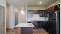 This thoughtfully designed 2-bedroom, 3-bathroom residence in Magnolia offers a functional and modern single-floor layout. The open-concept kitchen is a standout feature, showcasing rich dark cabinetry, granite countertops, black appliances, and a central island with pendant lighting overhead — seamlessly connecting to the living area, which is finished with hardwood-style flooring and carpeted transitions. Natural light flows generously through large windows fitted with blinds, complemented by ceiling fans in both the living area and bedrooms. Each bedroom features plush carpeting, a ceiling fan, and its own window with outdoor views. The primary bedroom connects directly to a private en-suite bathroom, which includes a granite-topped dark vanity, dual mirrors, and direct access to a spacious walk-in closet with built-in shelving. Additional bathrooms echo the same refined finishes, maintaining design consistency throughout. A dedicated laundry room provides in-unit washer and dryer hookups for added convenience. With six closets, two hallways, and a utility room, storage and practicality are prioritized throughout. The neutral color palette and cohesive design elements lend the space a clean, modern ambiance.