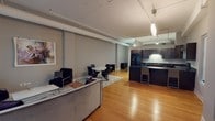 This single-floor urban residence showcases an industrial-modern aesthetic with an open-concept layout that seamlessly connects the living, dining, and kitchen areas. Soaring ceilings with exposed ductwork and track lighting lend a loft-like character throughout the main living space. Warm hardwood flooring extends across the entirety of the open area, providing a striking contrast to the cool gray walls.

The living area is anchored by large, floor-to-ceiling windows with sleek black frames, offering expansive city views and flooding the interior with natural light. The space comfortably accommodates both a seating arrangement and a separate dining area.

The kitchen is outfitted with rich, dark espresso cabinetry paired with black granite countertops and black appliances, including a refrigerator, range, microwave, and dishwasher. A peninsula with bar seating extends into the living area, reinforcing the open-plan design. Pendant lighting and under-cabinet illumination enhance functionality and ambiance.

Two bathrooms serve the unit. The full bathroom features dark tile flooring, a tub-shower combination with sliding glass doors and dark tile surround, and a dark vanity. The half bathroom offers a pedestal-style sink, tile flooring, and neutral wall tones, providing a compact and clean finish.