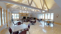 This single-floor community room is defined by its striking architectural design, most notably the soaring vaulted ceiling adorned with exposed white-painted timber trusses that create an open, cathedral-like atmosphere. Clerestory windows positioned along the upper roofline allow natural light to filter in from above, complementing the large windows lining the perimeter walls that frame views of the surrounding outdoor landscape.

The space is organized into two distinct zones. A casual lounge area is anchored by a full stone fireplace with a wood mantel, surrounded by upholstered sofas and armchairs arranged on a decorative rug, offering a comfortable gathering spot. The remainder of the room features multiple dining-style tables paired with burgundy chairs arranged on polished light-toned tile flooring, which reflects the abundant natural light and enhances the sense of spaciousness.

The neutral color palette of whites, light wood tones, and stone accents throughout creates a clean, welcoming ambiance suitable for a variety of community gatherings and events.