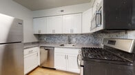 This single-floor unit offers a well-organized layout with distinct living, sleeping, and kitchen areas. The kitchen is a functional highlight, outfitted with crisp white shaker-style cabinetry, brushed nickel hardware, and a mosaic tile backsplash in soft gray tones. A full suite of stainless steel appliances includes a gas range, built-in microwave, dishwasher, and a full-size refrigerator. Light wood-tone flooring runs throughout the kitchen, complementing the gray laminate countertops and undermount sink. The living area is generously proportioned, featuring neutral-toned carpet and a sliding glass door that opens to a balcony with outdoor views of mature trees. Natural light fills the space through the expansive glass door and an additional window. The bedrooms are carpeted and offer ample room, with multiple doors suggesting closet access and connected spaces. The bathroom is fitted with a walk-in shower enclosed by sliding glass doors, featuring marble-style wall tile, a rain showerhead, and brushed nickel fixtures with a safety grab bar. Neutral tones throughout the unit create a clean, cohesive aesthetic that is both functional and move-in ready.