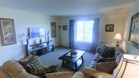 This single-story space features a living room designed for both comfort and functionality. The room is anchored by plush seating, including a sofa and a coordinating armchair, both upholstered in neutral tones and accented with patterned throw pillows. A dark-finished media console sits along one wall, topped with a flat-screen television and decorative accents, including vases and small ornamental pieces. Framed artwork on the walls adds visual interest to the light-colored interior. A large picture window, dressed with dark curtains, allows generous natural light to fill the space, creating a bright and airy atmosphere with views of outdoor greenery. The room is grounded by wall-to-wall carpeting in a warm neutral tone, contributing to the overall sense of comfort. A dark wood rectangular coffee table serves as the centerpiece of the seating arrangement, offering both style and practicality. The overall ambiance of the space is relaxed and inviting, with a balanced mix of functional furniture and decorative touches that create a welcoming environment suitable for everyday living.