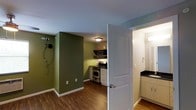 This single-floor space is thoughtfully designed to maximize functionality within a compact footprint. The main living area makes a strong visual statement with deep olive-green accent walls that contrast against crisp white trim and a cool grey ceiling, creating a cozy yet modern atmosphere. Rich, dark hardwood-style flooring runs throughout, adding warmth and continuity to the space.

The kitchen is tucked efficiently into the rear of the unit, featuring stainless steel appliances including a range and microwave, open shelving for accessible storage, and a brick accent wall that adds a touch of character and texture. White cabinetry provides ample storage, and a flush ceiling light keeps the area well-illuminated.

Accessible directly from the main living area, the bathroom is neatly appointed with a dark countertop vanity, light cabinetry, and a mirror, offering a clean and practical design. The layout of the space ensures smooth flow between the living area, kitchen, and bathroom, making everyday use intuitive and convenient. Overall, the unit balances utilitarian design with personalized touches that give it a distinct and inviting character.