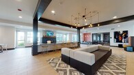 This expansive single-floor amenity space in Amherst, Massachusetts, is designed as a multifunctional social hub with a contemporary aesthetic. The open-plan layout is anchored by bold dark structural beams that contrast with light hardwood-style flooring and white ceilings, creating a modern industrial character throughout.

A central lounge area features a large sectional sofa set atop a geometric patterned rug, complemented by vibrant yellow accent chairs arranged near a sleek dark-tile fireplace with a wall-mounted television above a warm wood mantle. Multiple large windows allow natural light to fill the space and provide views of the surrounding exterior.

An adjacent bar-height counter with wood stools and a statement multi-bulb pendant chandelier serves as a gathering point, while a textured stone column adds a distinctive architectural detail. A bold red accent wall adds depth and visual contrast to the lounge area.

The game zone includes a full-size billiards table adorned by large-scale abstract artwork, as well as a ping-pong table. Upholstered bench seating in warm tones provides comfortable spectator areas. A glass-walled room visible from the main floor suggests additional private meeting or study space, rounding out this well-appointed communal environment.