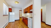This single-floor unit is characterized by consistent hardwood flooring that runs throughout the living areas, bedrooms, and kitchen, lending a warm and cohesive feel to the space. The living area is generously sized, featuring neutral cream-toned walls, dark wood baseboards, and a large window fitted with vertical blinds that allows ample natural light to fill the room. A baseboard heating system is present along the walls, ensuring year-round comfort.

The kitchen is compact yet functional, equipped with warm medium-toned wood cabinetry both above and below the counters, offering notable storage capacity. A double stainless steel sink sits beneath under-cabinet lighting, complemented by light-toned countertops. White appliances, including a range and refrigerator, complete the kitchen setup. The space connects directly to a hallway, providing easy flow throughout the unit.

Additional rooms include smaller rooms with windows fitted with horizontal blinds and baseboard heating, suitable for use as bedrooms or flexible spaces. The bathroom features classic white square tile surrounding a full soaking tub, a pedestal sink with chrome fixtures, a mirrored medicine cabinet above, and mosaic tile flooring, reflecting a traditional design aesthetic throughout.