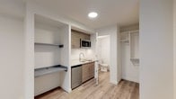 This thoughtfully designed single-floor unit offers a clean, modern aesthetic with a neutral color palette and light wood-style flooring that runs seamlessly throughout the space. The open living area is bright and airy, featuring a window with blinds that allows natural light to filter in, along with a wall-mounted thermostat and an electrical panel for added practicality.

The kitchenette is efficiently organized, featuring wood-toned cabinetry, a stainless steel sink with a modern arc faucet, an electric cooktop, an over-the-range stainless steel microwave, and a compact refrigerator — all set against crisp white countertops that enhance the clean, contemporary feel.

Adjacent to the kitchenette is a built-in shelving and desk nook, providing a dedicated workspace or storage area. A walk-in closet with built-in shelving and hanging rods offers well-organized storage solutions.

The bathroom is accessible directly from the main living area and includes a toilet and a glass-enclosed shower, maintaining the same clean white aesthetic. A hallway connects the various areas of the unit, contributing to a functional and efficient flow throughout the space.

The overall ambiance is fresh, minimalist, and move-in ready, making it a practical and well-appointed living space.