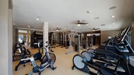 Located at 930 Jones Franklin Rd in Raleigh, NC, this comprehensive fitness center is thoughtfully designed to support a wide range of workout routines. The facility is organized into distinct zones, each tailored to a specific type of training. The main floor features a spacious cardio area lined with treadmills, ellipticals, and stationary bikes, including air resistance bikes, all positioned beneath ceiling fans that keep the environment comfortable. Large floor-to-ceiling windows along one wall offer direct views of an outdoor pool area, providing natural light and a refreshing backdrop during workouts. A dedicated strength training section houses a robust selection of equipment, including a power rack with barbells, multiple cable and selectorized machines by Precor, free weights, adjustable benches, and a heavy punching bag — catering to both novice and advanced lifters. A separate room, accessible through barn-style sliding doors, serves as a versatile studio space finished with hardwood-style flooring, full-length mirrored walls, ballet barre, a mounted flat-screen TV, and an assortment of functional training tools such as medicine balls and resistance bands. Recessed lighting throughout maintains a clean, modern aesthetic, while neutral taupe tones and white trim create a polished, welcoming atmosphere across the entire facility.