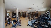 Located at 930 Jones Franklin Rd in Raleigh, NC, this comprehensive fitness center is thoughtfully designed to support a wide range of workout routines. The facility is organized into distinct zones, each tailored to a specific type of training. The main floor features a spacious cardio area lined with treadmills, ellipticals, and stationary bikes, including air resistance bikes, all positioned beneath ceiling fans that keep the environment comfortable. Large floor-to-ceiling windows along one wall offer direct views of an outdoor pool area, providing natural light and a refreshing backdrop during workouts. A dedicated strength training section houses a robust selection of equipment, including a power rack with barbells, multiple cable and selectorized machines by Precor, free weights, adjustable benches, and a heavy punching bag — catering to both novice and advanced lifters. A separate room, accessible through barn-style sliding doors, serves as a versatile studio space finished with hardwood-style flooring, full-length mirrored walls, ballet barre, a mounted flat-screen TV, and an assortment of functional training tools such as medicine balls and resistance bands. Recessed lighting throughout maintains a clean, modern aesthetic, while neutral taupe tones and white trim create a polished, welcoming atmosphere across the entire facility.