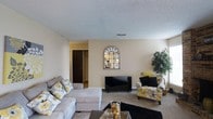 This single-floor residence offers two bedrooms and two bathrooms within a well-proportioned layout. The living room serves as the central gathering space, featuring wall-to-wall carpeting that adds warmth underfoot and a textured ceiling that gives the room subtle character. Neutral beige walls provide a versatile backdrop, allowing for a variety of décor styles. A dedicated media wall offers space for a flat-screen television, complemented by a decorative arched mirror that adds visual interest and depth to the room. The living area comfortably accommodates a sectional sofa and accent seating, demonstrating the generous scale of the space. A hallway visible from the living room leads to additional areas of the home, including what appears to be a spacious closet with built-in shelving, highlighting the storage capacity throughout. Ceiling vents suggest a central HVAC system for year-round climate comfort. The overall ambiance is bright and welcoming, with a neutral color palette that creates a calm and livable atmosphere throughout this well-sized single-story home.