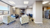 Located at 95 Cambridge St in Cambridge, Ontario, this single-floor common lounge offers a well-appointed, multi-functional layout designed to accommodate a variety of social and recreational activities. The space is carpeted throughout with a patterned tile-style carpet that adds texture and warmth underfoot.

The room is organized into distinct functional zones. A lounge area anchors one end of the space, featuring a stone-faced fireplace with a mounted flat-screen television above it, flanked by navy blue leather sofas and patterned accent chairs — creating a cozy, inviting atmosphere. Adjacent to this is a dining or gathering area with a round table and upholstered chairs, complemented by a large decorative wall clock and views through a bank of windows that draw in generous natural light and offer outdoor sightlines.

A compact kitchenette is also integrated into the space, equipped with wooden cabinetry, a stainless steel refrigerator, microwave, and a sink — supporting light refreshment and catering needs. Additional seating arrangements and a console with decorative accents provide further flexibility throughout the room.

The drop ceiling with recessed panel lighting ensures consistent, even illumination throughout. A separate entry door with emergency signage provides practical access. Overall, the space presents a comfortable, community-oriented environment suited for lounging, dining, or socializing.