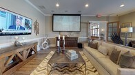 This single-floor amenity space is designed for entertainment and socializing, combining multiple functional zones within one cohesive, open layout. The interior is finished with warm hardwood-style flooring, crown molding, and neutral-toned walls accented by wainscoting, creating a polished yet welcoming atmosphere throughout.

The main lounge area serves as a home theater, anchored by a large projection screen and ceiling-mounted audio system. Comfortable seating, including a generous sofa and upholstered chairs, is arranged around a substantial wood-and-metal coffee table atop a chevron-patterned area rug. Decorative wall art and sculptural accents add visual interest.

Adjacent to the theater area, a billiards table occupies a generously sized open zone, complemented by additional seating arrangements with televisions mounted on the walls, allowing for multiple simultaneous activities.

A fully equipped kitchen features light wood cabinetry extending to the ceiling, marble-look countertops, a large island with bar seating, and a wall-mounted television. Geometric pendant lighting adds a contemporary touch overhead.

A quieter lounge nook near large windows offers natural light, leather seating, and a dining table with chairs, providing a more intimate setting within the broader space. Recessed lighting throughout ensures consistent illumination across all zones.