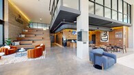 The Mercer lobby and amenity space at 2078 19th St in Denver, Colorado, is a thoughtfully designed communal hub that seamlessly blends contemporary architecture with warm, inviting interiors. The space is anchored by dramatic floor-to-ceiling curtain-wall glazing along the street-facing façade, flooding the interior with abundant natural light while offering open views of the surrounding Denver streetscape.

Soaring double-height ceilings create an expansive, airy atmosphere, with exposed industrial ductwork adding a modern edge against the polished concrete floors and rich warm-toned wood wall paneling. A sculptural staircase with cantilevered wood-clad steps serves as a striking architectural focal point, leading to an upper mezzanine level enclosed by steel-framed glass railings.

The layout is organized into distinct functional zones. A welcoming reception area features a distinctive fluted green desk paired with plush rust-orange lounge chairs arranged on textured area rugs. A café and bar counter with warm wood cabinetry and pendant lighting offers a casual gathering point. Further into the space, communal dining and coworking tables with mixed seating—including navy upholstered chairs—provide flexible work and social settings. The overall ambiance is sophisticated yet approachable, combining industrial and residential design elements cohesively.