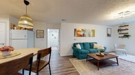 This single-floor unit offers a thoughtfully designed layout that balances comfort and functionality across its key living spaces. The living room serves as the heart of the home, featuring durable wood-look flooring, a textured ceiling, and a modern flush-mount light fixture. A wall-mounted television, floating wooden shelves adorned with trailing plants, and a large window that brings in natural light complete the inviting atmosphere. A teal sofa, accent chair, and mid-century coffee table add warmth and character to the space. The open layout connects seamlessly to a dining area, making the space feel open and cohesive. The kitchen is equipped with white cabinetry, a full suite of white appliances including a refrigerator, range, microwave, and dishwasher, along with warm-toned countertops and a skylight-style ceiling fixture that brightens the workspace. The bedroom offers plush carpeting, a generously sized window with garden views, and ample room for a full bedroom suite. The bathroom is clean and functional, featuring a shower-tub combination with a curtain, a vanity, and a neutral color palette. A dedicated laundry area with washer, tile flooring, and utility storage rounds out this practical and comfortable home.