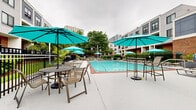 Located at 105 Lighthouse Terrace in Edgewater, New Jersey, this outdoor amenity area offers a well-appointed residential pool and recreation space designed for relaxation and leisure. The centerpiece is a generously sized swimming pool with clear, blue water, framed by a classic brick coping border and surrounded by an expansive concrete sundeck. The deck is well-furnished with an abundance of lounge chairs and chaise lounges, providing ample space for sunbathing and outdoor enjoyment. Vibrant teal patio umbrellas are stationed throughout the area, offering shaded seating options at both high-top and standard dining tables. A dedicated outdoor lounge zone features neutral-toned sectional sofas with decorative pillows, creating a comfortable social gathering space. A poolside support structure with a standing seam metal roof houses pool amenities and is accented by a nautical compass wall decoration, nodding to the property's lighthouse-inspired identity. The entire pool area is enclosed by a black iron fence, ensuring privacy and security. Mature trees and tropical plantings soften the perimeter, adding a lush, resort-like atmosphere. The multi-story residential building backdrop, finished in a contemporary mix of gray paneling and brick, provides an attractive architectural frame for this outdoor retreat.