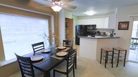 This single-floor space showcases an open-concept layout that seamlessly connects the dining area and kitchen, creating a cohesive and functional living environment. The dining area is anchored by a dark-stained wood table paired with matching chairs, positioned beneath a ceiling fan with a built-in light fixture. A large window with horizontal blinds fills the space with natural light, enhancing the warm, inviting atmosphere. Adjacent built-in shelving provides convenient storage and display options.

The kitchen features crisp white upper cabinetry contrasted against dark countertops, creating a classic two-tone aesthetic. Black appliances, including a refrigerator, microwave, and range, complement the design. A peninsula-style breakfast bar extends into the open space, offering additional seating via a wooden barstool and serving as a natural divider between the kitchen and dining areas. A double sink with a window-facing position adds functionality. The overall color palette, featuring warm tan and soft green walls, contributes to a comfortable and welcoming ambiance throughout the space.