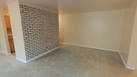 This single-floor space blends classic architectural charm with a warm, understated aesthetic. The most striking feature is a full exposed brick accent wall, painted in a light whitewash finish that adds texture and visual interest while maintaining a clean, bright feel. The brick wall stands in pleasing contrast to the smooth, neutral-toned walls that complete the room, which are finished with crisp white baseboards for a tidy, polished look. The floors are covered in plush, neutral-toned carpet that adds warmth and comfort underfoot, contributing to the overall cozy ambiance of the space. The room is generously proportioned, offering an open and versatile layout with ample room for a variety of furniture arrangements. The neutral color palette throughout — from the beige walls to the soft carpeting — creates a calm, inviting atmosphere that serves as an excellent backdrop for personalization. The space is well-suited for use as a living area or multi-purpose room. Its single-floor layout ensures easy accessibility and a seamless flow throughout. Overall, this space combines a touch of rustic character through its brick detailing with a simple, functional design that appeals to a wide range of tastes.