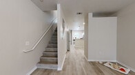 This two-story home presents a clean, modern aesthetic defined by light wood-style flooring and neutral warm-beige walls throughout. The main level opens into a welcoming entryway where a staircase with a white railing leads to the upper floor, while a hallway guides residents toward the living and kitchen areas. The kitchen is well-appointed with white cabinetry, light quartz-style countertops, stainless steel appliances including an electric range, over-the-range microwave, and dishwasher, along with a double-basin sink positioned beneath a window that brings in natural light and outdoor views. Adjacent to the kitchen, a dedicated laundry closet houses a full-size washer and dryer with overhead shelving for storage. A bathroom on the main level features a tub-shower combination with a curtain, a vanity with a light countertop, and a large mirror. Additional rooms across both floors offer flexible spaces suitable for bedrooms, a home office, or exercise areas, each featuring the consistent light flooring and neutral walls. Windows fitted with horizontal blinds allow natural light while maintaining privacy, contributing to the bright, airy ambiance found throughout the home.