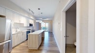 Located at 200 Daytona Blvd in Daytona Beach, FL, this thoughtfully designed single-story residence offers 1 bedroom and 2 bathrooms within a bright, contemporary layout. The open-concept living area seamlessly connects the kitchen, dining, and living room, creating an airy and functional space bathed in natural light from a sliding glass door that leads to a private patio. From the patio, residents enjoy scenic views of lush palm-lined grounds and the surrounding Daytona Beach community. The modern kitchen is a standout feature, boasting a large center island with a light wood finish, crisp white cabinetry reaching to the ceiling, stainless steel appliances including a refrigerator, range, microwave, and dishwasher, and contemporary circular pendant lighting. Light wood-tone flooring flows throughout the main living areas, complementing the neutral warm-toned walls. The spacious primary bedroom includes a generous closet and access to a full bathroom featuring a glass-enclosed walk-in shower and tile flooring. A second bathroom offers a pedestal sink and an elegant oval mirror, ideal for guests. Additional conveniences include a dedicated laundry area, utility room, and two closets, making this residence as practical as it is stylish.