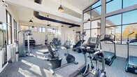Located on Tryon Road in Raleigh, NC, this single-floor fitness center is designed to deliver a premium workout experience through thoughtful architecture and a well-curated equipment selection. The space is defined by dramatically high ceilings with exposed ductwork that adds an industrial aesthetic, complemented by sleek pendant lighting and ceiling fans that maintain comfort throughout. Two distinct zones make up the facility. The cardio area faces an expansive wall of floor-to-ceiling steel-framed windows, flooding the space with natural light and offering outdoor views of the surrounding community, including a pool area. This zone houses a row of TRUE brand treadmills, ellipticals, recumbent bikes, and stair climbers, each equipped with individual screens for entertainment. The strength training zone occupies a separate wing with its own clerestory windows along the upper walls. It features a multi-station cable and resistance machine, a full dumbbell rack with an adjustable bench, and two dedicated TRX Training Zone rigs stocked with kettlebells, resistance bands, and suspension trainers. Full-length mirrors line portions of the walls, enhancing the sense of space. Patterned carpet tile flooring throughout ties both zones together, creating a cohesive and functional environment.