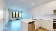 This well-designed 1-bedroom, 1-bathroom unit offers a thoughtfully planned layout that maximizes functionality and natural light throughout. The open-plan kitchen and living area serves as the heart of the space, featuring sleek white cabinetry extending to the ceiling, a white quartz peninsula with an undermount sink, and a full suite of stainless steel appliances including a refrigerator, range, microwave, and dishwasher. Light hardwood flooring runs seamlessly through the main living areas, complementing the clean, contemporary aesthetic. Large black-framed windows in the living area invite natural light and offer views of the surrounding neighborhood. The bedroom is generously proportioned, featuring the same hardwood flooring, large windows with outdoor views, a wall-unit HVAC system, and a spacious built-in closet with shelving and hanging rails. The bathroom is finished with a light wood vanity, white quartz countertop, an undermount sink, and a large mirror, lending a warm yet modern feel. Louvered closet doors near the entryway provide additional storage. Throughout the unit, a neutral white and warm beige palette, recessed lighting, and consistent finishes create a bright, cohesive, and move-in-ready living environment.