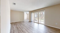 This single-floor residence is designed with a clean, functional aesthetic that emphasizes natural light and open space. Light-toned wood-look flooring runs consistently throughout the living areas and bedrooms, creating a cohesive and easy-to-maintain foundation. The walls are finished in a neutral warm beige, complemented by white baseboards and textured popcorn ceilings.

The main living area is generously proportioned and benefits from large sliding glass doors that open to an outdoor view of mature trees and greenery, filling the space with abundant natural light. A ceiling fan is visible in an adjacent area, indicating a connected secondary living or dining zone that flows naturally from the main room.

The kitchen features white shaker-style cabinetry with brushed silver hardware, light countertops, a stainless steel sink, and black appliances including a range and over-the-range microwave. The layout offers ample counter and storage space in a functional configuration.

The bedrooms are well-sized, each with wood-look flooring and windows that introduce natural light. The bathrooms are finished with white shaker vanity cabinetry, light countertops, large mirrors, and tiled tub-shower combinations with chrome fixtures, presenting a clean and practical design throughout.