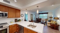 Unit #104 at 2615 Con non Rd in Carlsbad is a well-appointed single-floor residence designed for comfortable everyday living. The open-concept layout connects the kitchen, dining, and living areas into one cohesive space, promoting a sense of openness and ease of movement throughout.

The living and dining area features wood-look flooring, crown molding, and a large window with natural light illuminating the room. The combined space comfortably accommodates both a dining set and a full living room arrangement, anchored by a glass-top oval coffee table and a plush sofa.

The kitchen is a functional highlight, featuring warm wood cabinetry, white quartz countertops, stainless steel appliances including a French door refrigerator and dishwasher, pendant lighting over the peninsula, and a built-in sink. Bar-height seating along the peninsula adds additional dining flexibility. An arched doorway connects the kitchen to adjacent areas, reinforcing the home's traditional architectural detailing.

The bedroom is a quiet, carpeted retreat with a window offering natural light. The full bathroom includes wood cabinetry with a white countertop vanity, wood-look flooring, and a shower-tub combination equipped with accessibility grab bars, reflecting a thoughtfully designed layout.