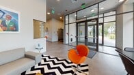 Located on Melnea Cass Boulevard in Boston's Roxbury neighborhood, this single-floor lobby space presents a thoughtfully designed, contemporary interior that balances functionality with artistic expression. The entry area features floor-to-ceiling glass curtain walls and double doors, flooding the space with natural light and offering clear views of the surrounding streetscape and greenery beyond.

The lounge area is anchored by a sleek gray sofa positioned atop a bold black-and-white chevron area rug, accompanied by a sculptural orange accent chair that adds a vibrant pop of color. A large, colorful figurative painting displayed prominently above the sofa serves as a striking focal point, contributing cultural depth to the space. Built-in shelving and cabinetry in light wood tones provide functional storage throughout.

A conference or meeting area features a substantial dark wood table surrounded by upholstered chairs, positioned directly in front of expansive grid-paned windows that frame lush outdoor views. An accent wall clad in light wood-toned paneling hosts a wall-mounted flat-screen television, completing the modern, polished aesthetic. Light tile flooring ties the areas together, enhancing the open, airy atmosphere throughout.