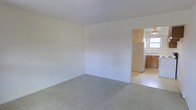 This single-floor space features a straightforward, functional layout with a clear separation between the living area and kitchen. The main living area is finished with wall-to-wall neutral-toned carpeting, which adds a sense of warmth underfoot. The walls are painted in a clean, off-white tone throughout, contributing to a bright and open atmosphere. The room is generously sized, offering a versatile space suitable for a variety of furniture arrangements. Ceilings are standard height, maintaining a comfortable, enclosed feel.

An open doorway connects the living area to the kitchen, which transitions to hard flooring. The kitchen features warm-toned wooden cabinetry providing ample storage, a white refrigerator, and a sink positioned beneath a window that allows natural light to filter into the space. The window offers the kitchen a welcoming brightness. The overall design is practical and unfussy, prioritizing usability. The neutral color palette carried throughout both spaces creates a cohesive and clean aesthetic, making it easy to personalize with furnishings and décor.
