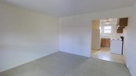 This single-floor space features a straightforward, functional layout with a clear separation between the living area and kitchen. The main living area is finished with wall-to-wall neutral-toned carpeting, which adds a sense of warmth underfoot. The walls are painted in a clean, off-white tone throughout, contributing to a bright and open atmosphere. The room is generously sized, offering a versatile space suitable for a variety of furniture arrangements. Ceilings are standard height, maintaining a comfortable, enclosed feel.

An open doorway connects the living area to the kitchen, which transitions to hard flooring. The kitchen features warm-toned wooden cabinetry providing ample storage, a white refrigerator, and a sink positioned beneath a window that allows natural light to filter into the space. The window offers the kitchen a welcoming brightness. The overall design is practical and unfussy, prioritizing usability. The neutral color palette carried throughout both spaces creates a cohesive and clean aesthetic, making it easy to personalize with furnishings and décor.