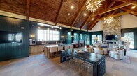This single-story community space showcases a striking open-concept design anchored by soaring vaulted ceilings clad in warm knotty wood planks and supported by exposed timber beams. The expansive main area is unified by large-format stone tile flooring that extends throughout, lending a refined yet rustic character to the space. Deep teal shiplap paneling wraps the walls, creating a bold, cohesive color palette that is both inviting and sophisticated.

A dramatic floor-to-ceiling natural stone fireplace serves as a focal point in the lounge area, topped by a wall-mounted television and flanked by comfortable seating. Large glass-paned doors and multi-pane windows flood the interior with natural light and offer views of an outdoor pool area. Statement starburst chandeliers add a modern, sculptural contrast to the rustic architectural elements above.

The space is thoughtfully zoned to accommodate multiple uses. A kitchen bar with a large island and counter seating supports casual dining and entertaining, while dark wood communal tables and mixed seating arrangements define flexible gathering and work areas. A built-in workstation with dual monitors provides a dedicated workspace. An entry corridor with teal-painted shiplap walls connects the main hall, completing this well-appointed, multifunctional space.