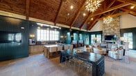 This single-story community space showcases a striking open-concept design anchored by soaring vaulted ceilings clad in warm knotty wood planks and supported by exposed timber beams. The expansive main area is unified by large-format stone tile flooring that extends throughout, lending a refined yet rustic character to the space. Deep teal shiplap paneling wraps the walls, creating a bold, cohesive color palette that is both inviting and sophisticated.

A dramatic floor-to-ceiling natural stone fireplace serves as a focal point in the lounge area, topped by a wall-mounted television and flanked by comfortable seating. Large glass-paned doors and multi-pane windows flood the interior with natural light and offer views of an outdoor pool area. Statement starburst chandeliers add a modern, sculptural contrast to the rustic architectural elements above.

The space is thoughtfully zoned to accommodate multiple uses. A kitchen bar with a large island and counter seating supports casual dining and entertaining, while dark wood communal tables and mixed seating arrangements define flexible gathering and work areas. A built-in workstation with dual monitors provides a dedicated workspace. An entry corridor with teal-painted shiplap walls connects the main hall, completing this well-appointed, multifunctional space.