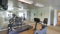This fitness center offers a dedicated space for exercise and wellness, thoughtfully designed to accommodate a variety of workout routines. The room is outfitted with an array of cardio equipment, including treadmills and a stationary bike, providing ample options for cardiovascular training. Resistance and strength training equipment is also available, rounding out the facility's offerings for a full-body workout.

Floor-to-ceiling mirrors line the walls, creating a sense of openness and allowing users to monitor their form during exercise. Overhead fluorescent lighting ensures the space is consistently well-lit throughout the day. The neutral-toned carpeted flooring adds a degree of comfort underfoot, while the clean, functional layout keeps the space organized and easy to navigate.

A window provides a connection to natural light, further brightening the room. Wall-mounted accessories and storage elements contribute to the facility's practical design. The overall ambiance is clean, focused, and purpose-driven, making this fitness center a convenient and functional amenity for those looking to maintain an active lifestyle.