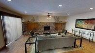 This single-floor living space is anchored by a commanding, full-width brick fireplace wall that serves as the room's defining architectural feature. The fireplace is framed by built-in wooden cabinetry with a continuous wood mantel shelf spanning the entire wall, creating a cohesive and warm focal point. Decorative wall art flanks both sides of the mantel, adding personality to the space.

The room is finished with rich hardwood-style flooring that complements the warm tones of the brick and wood cabinetry. A textured popcorn ceiling overhead features strategically placed recessed lighting alongside a central ceiling fan, ensuring both functional illumination and comfortable air circulation throughout the space.

The living area is generously sized, comfortably accommodating a large sectional sofa, paired lounge chairs, and a metal-framed coffee table arranged over a layered area rug. The neutral wall tones allow the brick feature wall to remain the visual centerpiece, while the overall layout promotes an inviting, lived-in ambiance ideal for relaxed gatherings or everyday comfort.