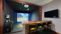 Located at 20200 Poplar Creek Pkwy in Brookfield, Wisconsin, this indoor golf simulator suite offers a well-appointed, self-contained experience designed for both practice and entertainment. The space is accessed through a corridor framed by rich wood-trimmed glass panel doors with craftsman-style detailing, setting a refined tone from the moment of entry.

At the heart of the suite is a Full Swing golf simulator, complete with a dedicated hitting bay featuring padded walls and a synthetic turf hitting mat. A ceiling-mounted projector delivers a vivid, large-format display of photorealistic golf courses, while ambient blue LED accent lighting along the base of the simulator enclosure adds a modern, immersive atmosphere.

Adjacent to the hitting bay, a generously sized counter with a live-edge wood top provides a comfortable viewing and lounging area, accompanied by bright yellow-green chairs that add a pop of color. A wall-mounted flat-screen TV and a decorative sconce light the lounge side of the room, and framed golf artwork lines the walls. A compact equipment cabinet provides organized storage nearby.

The flooring throughout features durable charcoal-toned carpet tiles, complemented by warm wood baseboards and trim that unify the design. The overall ambiance is that of a polished, professionally outfitted simulator bay ideal for year-round golf practice or group entertainment.