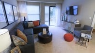 This single-floor living space features a well-appointed living room that serves as its central gathering area. Large windows fitted with white blinds and sheer curtains allow abundant natural light to fill the room, while a sliding glass door opens to an outdoor area, offering views of surrounding greenery and further enhancing the sense of openness.

The living area is designed for both comfort and style. A spacious sectional sofa in a deep charcoal tone anchors the space, accented with teal, rust, and patterned throw pillows that introduce a vibrant color palette. A round leather ottoman positioned at the center doubles as a coffee table, adding a functional yet contemporary focal point. A knitted rust-colored pouf provides additional seating and visual interest.

Along one wall, a multi-toned media console in a patchwork finish supports a wall-mounted flat-screen television, complemented by decorative accents including tall vases and sculptural pieces. The neutral carpeted flooring ties the room together, reflecting the warm ambient lighting. Overall, the space presents a balanced combination of modern design and inviting comfort.