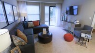 This single-floor living space features a well-appointed living room that serves as its central gathering area. Large windows fitted with white blinds and sheer curtains allow abundant natural light to fill the room, while a sliding glass door opens to an outdoor area, offering views of surrounding greenery and further enhancing the sense of openness.

The living area is designed for both comfort and style. A spacious sectional sofa in a deep charcoal tone anchors the space, accented with teal, rust, and patterned throw pillows that introduce a vibrant color palette. A round leather ottoman positioned at the center doubles as a coffee table, adding a functional yet contemporary focal point. A knitted rust-colored pouf provides additional seating and visual interest.

Along one wall, a multi-toned media console in a patchwork finish supports a wall-mounted flat-screen television, complemented by decorative accents including tall vases and sculptural pieces. The neutral carpeted flooring ties the room together, reflecting the warm ambient lighting. Overall, the space presents a balanced combination of modern design and inviting comfort.