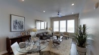 This single-floor space showcases a thoughtfully designed open-plan layout that seamlessly connects the dining and living areas. The living room is anchored by a plush, cream-toned area rug and furnished with a dark leather sofa and complementary neutral-toned accent chairs arranged around a sleek glass-and-chrome coffee table. A ceiling fan with an integrated light fixture serves as a functional focal point overhead, while recessed lighting throughout ensures consistent, even illumination. Large windows fitted with horizontal blinds allow an abundance of natural light to fill the space, creating a bright and welcoming ambiance. A large framed mirror on the wall further amplifies the sense of openness. The dining area features a round glass-top table with chrome detailing, elegantly set with formal place settings, wine glasses, and decorative accents. A dark wood sideboard along the wall displays framed artwork and personal photographs. Warm hardwood-style flooring runs throughout, grounding the neutral color palette of grays, creams, and browns. The overall atmosphere is modern, sophisticated, and inviting, with carefully selected furnishings and décor contributing to a polished, cohesive aesthetic.