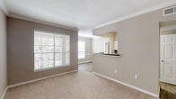 This single-story, one-bedroom, one-bathroom residence offers a thoughtfully designed open floor plan with neutral warm taupe walls and white crown molding throughout the main living areas. The spacious living room is carpeted and filled with natural light from multiple large windows fitted with horizontal blinds, creating a bright and airy atmosphere. A ceiling fan overhead enhances comfort and airflow.

The kitchen connects seamlessly to the living area via a pass-through opening with a dark granite-style countertop bar, offering both functional workspace and a visual connection between rooms. The kitchen is equipped with white cabinetry, dark granite-style countertops, a black electric range with coil burners, and a matching over-the-range microwave, complemented by wood-look flooring.

The full bathroom features a dual-sink vanity with dark granite-style countertops, white cabinetry, wood-look flooring, and a well-lit mirror above. A louvered closet door conceals an in-unit washer and dryer, adding notable convenience.

The bedroom is generously sized with plush carpet, a ceiling fan, and a walk-in closet. French doors in one area open to the exterior, allowing additional natural light and outdoor access throughout the residence.