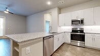This thoughtfully designed single-floor residence offers a clean, contemporary aesthetic throughout, highlighted by warm wood-look plank flooring and a neutral color palette that creates a bright, welcoming atmosphere.

The kitchen is a standout feature, offering generous U-shaped cabinetry in crisp white shaker-style with sleek bar pulls, granite countertops, a subway tile backsplash, and a full suite of stainless steel appliances including a refrigerator, range, over-the-range microwave, and dishwasher. A peninsula provides additional counter space and casual seating.

Adjacent to the kitchen, a dedicated built-in bar area features granite countertops, white cabinetry, and open wine rack shelving above — an ideal space for entertaining.

The living areas are spacious and airy, with ceiling fans, large windows, and a glass door leading to the outdoors, flooding the space with natural light and offering views of the surrounding exterior.

Bedrooms are well-proportioned with large windows and the same wood-look flooring found throughout. The primary bathroom is elegantly appointed with a granite-top white vanity, a framed mirror, and direct access to a spacious walk-in closet with multi-tiered shelving and hanging rods. The overall design balances functionality and style seamlessly.
