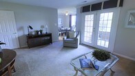 This single-floor residence offers a welcoming and well-lit interior characterized by a seamless open-concept layout. The living area is finished with soft wall-to-wall carpeting and neutral gray walls that create a calm, versatile backdrop for furnishings. A ceiling fan overhead adds both function and a subtle design touch. The space is anchored by a glass-top coffee table and complemented by a rich dark wood media console along the wall, providing both storage and display space. A comfortable accent chair with a decorative pillow adds a cozy focal point to the room. Large French doors with white grid panes flood the living area with natural light and provide direct access to an outdoor patio or deck area, where lush greenery is visible beyond the glass. The open layout flows naturally into a dedicated dining area, which features a classic dining table and chairs set beneath a window dressed with dark valances and curtains. The consistent flooring and color palette throughout both spaces reinforce a cohesive, unified design. Together, these areas create a functional and inviting interior that balances comfort and style in a bright, open environment.