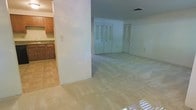 This single-floor space presents a practical and open layout designed for comfortable everyday living. The main living area is finished with wall-to-wall carpet, creating a warm and inviting atmosphere, while transition areas feature tile flooring that adds a clean contrast. A partial wall divides the living space from the adjacent kitchen, which is outfitted with warm wood-toned cabinetry and countertops, providing ample storage and workspace. The kitchen's tile flooring complements the cabinetry and connects seamlessly to the main living area. Natural light filters into the space, contributing to a bright and airy feel throughout. The far end of the living area features a set of double closet doors alongside an additional single door, offering generous built-in storage options. The walls are finished in neutral tones, providing a versatile backdrop that suits a variety of interior styles. Overhead, standard ceiling height with a centrally located ventilation register ensures consistent airflow throughout the space. Overall, the layout is efficient and adaptable, making it well-suited for a range of uses.