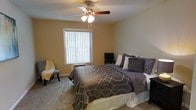 This single-floor space features a well-appointed bedroom that serves as a comfortable retreat. The room is finished with wall-to-wall carpeting in a neutral tone, contributing to a warm and inviting atmosphere. Walls painted in a soft beige provide a calming backdrop that complements the room's understated decor.

A large window fitted with vertical blinds allows natural light to filter into the space while offering adjustable privacy. Positioned centrally on the ceiling, a multi-blade ceiling fan with an integrated light fixture provides both ambient lighting and airflow, enhancing year-round comfort.

The bedroom offers enough space to accommodate a generously sized bed dressed in layered grey bedding with a geometric pattern, alongside a bedside table and a small media setup. A cushioned accent chair positioned near the window adds a functional seating option. The overall layout is practical and efficient, making good use of the available floor space without feeling crowded. The neutral tones, soft flooring, and natural light combine to create a relaxed and livable environment.