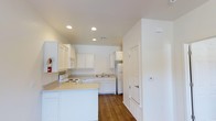 This single-floor 1-bedroom, 1-bathroom residence delivers a functional and comfortable living environment with a clean, modern aesthetic throughout. The kitchen serves as a central highlight of the home, featuring crisp white cabinetry that extends to the ceiling, light-toned countertops, and a full suite of white appliances including a range, dishwasher, and refrigerator. A kitchen island provides additional prep space and enhances the room's utility. Recessed lighting illuminates the kitchen evenly, complementing the warm hardwood-style flooring that runs throughout the space. The neutral white walls and ceiling create a bright, open atmosphere that makes the interior feel airy and inviting. A thermostat is conveniently mounted near the kitchen entry, indicating climate control features within the unit. The layout is efficiently organized to maximize the use of available space, with clear sightlines from the kitchen into the adjoining living areas. Overall, this residence offers a tidy and practical floor plan suited for comfortable everyday living, with cohesive finishes that lend a fresh, well-maintained character to the home.