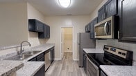 This single-floor, 2-bedroom, 2-bathroom residence offers a thoughtfully designed layout with modern finishes and a welcoming ambiance. The galley-style kitchen is a standout feature, showcasing dark slate cabinetry with sleek hardware, granite-style countertops, and a full suite of stainless steel appliances including a refrigerator, microwave, and range. Wood-look plank flooring flows throughout the main living areas, lending a clean and cohesive aesthetic. A peninsula with a granite countertop connects the kitchen to the open living and dining area, which is anchored by a fireplace and ceiling fan, and benefits from natural light through multiple windows. A dedicated laundry room is conveniently accessible directly from the kitchen. Both bedrooms are generously sized, featuring plush carpet, ceiling fans, and windows that bring in natural light. Each bathroom is well-equipped, offering a soaking tub with white tile surround, and one bathroom includes a granite-topped vanity with dark cabinetry. The overall design balances practicality and comfort, making this a functional and attractive living space.